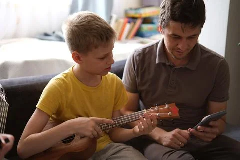 Father using smartphone teaching son to play ukulele at home Stock Photos