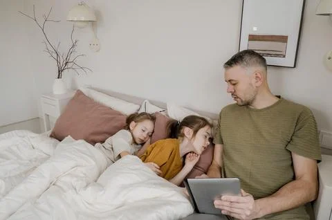 A father is using a tablet while his two daughters are sleeping peacefully in Stock Photos