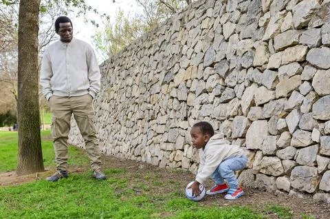 Father watching son playing soccer in park by stone wall Stock Photos