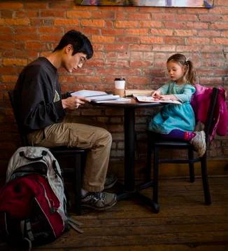 Father working while daughter reading book while sitting against brick wall at Stock Photos