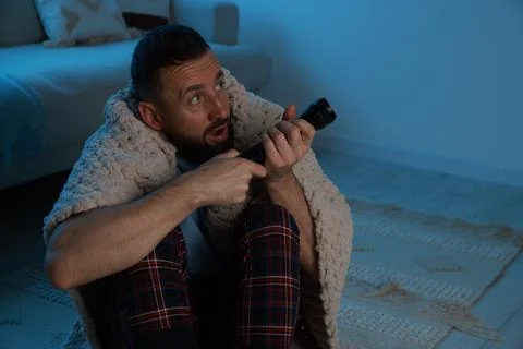 Fear of darkness. Scared man with blanket and flashlight indoors at night, .. Stock Photos