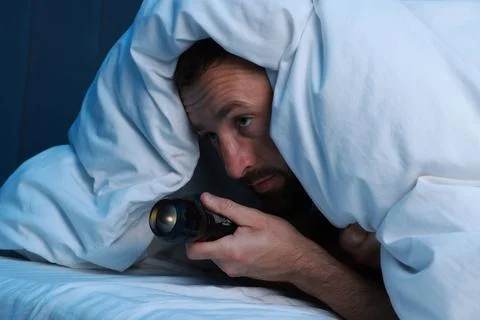 Fear of darkness. Scared man with blanket and flashlight on bed in room at .. Stockfoto's