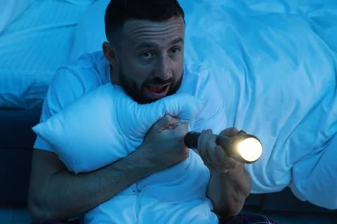 Fear of darkness. Scared man with flashlight hugging pillow in bedroom at nig Fotos de archivo