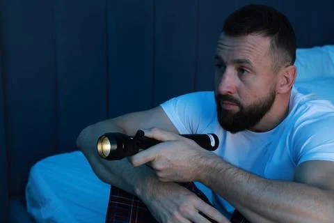 Fear of darkness. Scared man with flashlight sitting on bed in room at night Stockfoto's