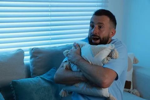 Fear of darkness. Scared man hugging pillow on couch in dark room Stock Photos