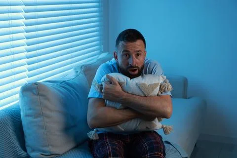 Fear of darkness. Scared man hugging pillow on couch in dark room Stock Photos