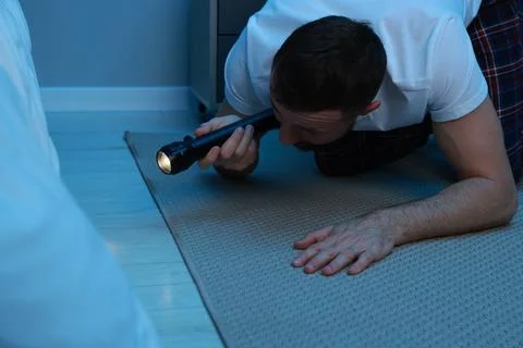 Fear of darkness. Scared man looking under bed with flashlight indoors at nig Stock Photos