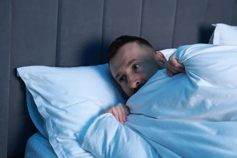 Fear of darkness. Scared man lying on bed in room at night 스톡 사진