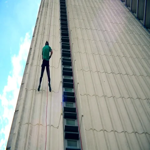 Fearless man descend down with cord on skyscraper wall. Blue cloudy sky Stock Footage 69473001