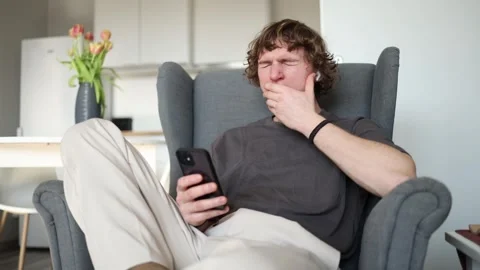 Feeling sleepy A young man sitting in a chair with a phone in his hand, yawning  Stock Footage 302634141