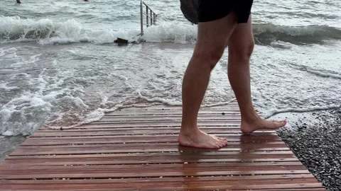 The feet of a man cross the frame against a wooden walkway going to the sea Stock Footage 250508063