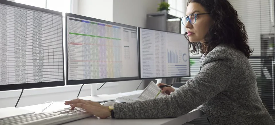 Female accountant auditing data on multiple monitors in modern office workspace Stock Photos