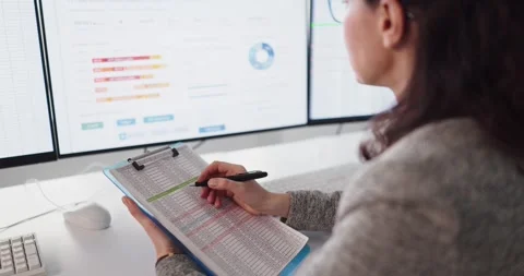 Female accountant checking spreadsheet on clipboard near monitors in modern Stock Footage 330941500