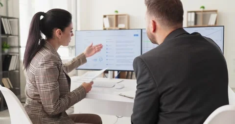 Female analyst presenting data to colleague on dual monitors in modern office Stock Footage 337619365