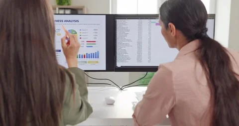 Female analysts reviewing financial data on dual monitors during office teamwork Stock Footage 344006222