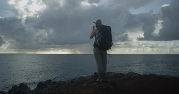Female backpacker climbing on hill towards a view of a stormy sea Stock Footage 86110775