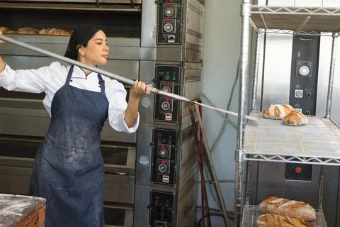 Female baker in chef jacket, floured blue apron handling loaves with peel at Stock Photos