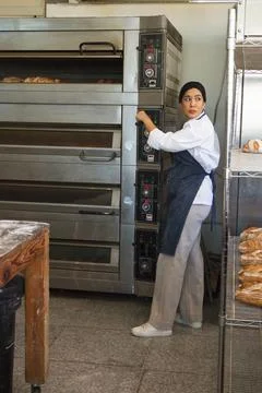 Female baker operating multi-deck oven controls while checking loaves on Stock Photos