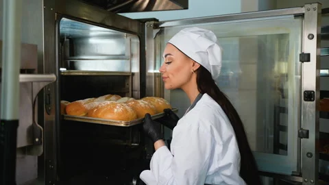 Female baker pulling hot bread tray from oven Stock Footage 309890397
