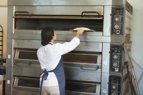 Female baker wearing chef jacket and apron placing dough using peel at Stock Photos