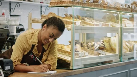 Female baker writing in the notebook while working at her store Stock Footage 77178518