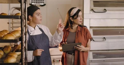 Female bakery team at racks, baker in chef coat pointing while manager using Stock Footage 338064548