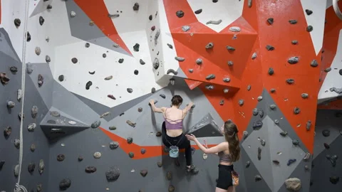 Female boulderer working on challenging route in gym while friend observes and Stock Footage 346542730