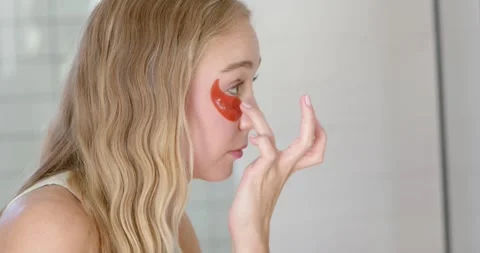 Female checking mirror and adjusting red under-eye gel patch at bathroom vanity, Video stock 335987567