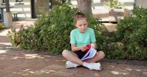 Female child in mint tee at school under tree, practicing reading, turning pages Stock Footage 340773477