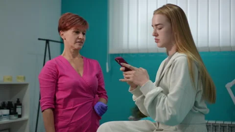 Female client using smartphone while cosmetologist explains procedure in clinic Stock Footage 311363763
