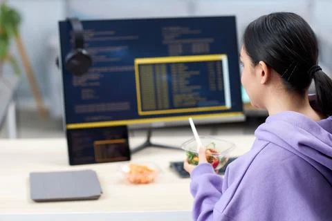 Female computer programmer eating takeout lunch at workplace while writing code Foto stock