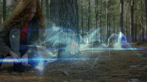Female designer crouching in pine forest clearing, interacting with blue energy Stock Footage 308185526