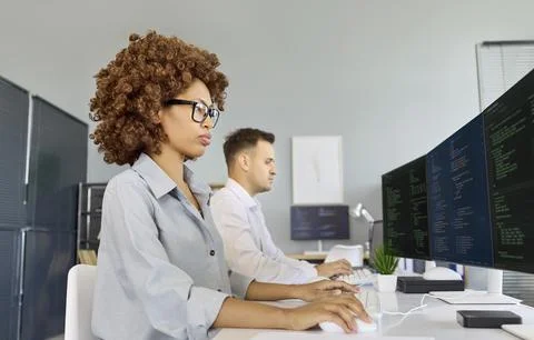 Female developer coding at multi monitor workstation with colleague in office Stock Photos