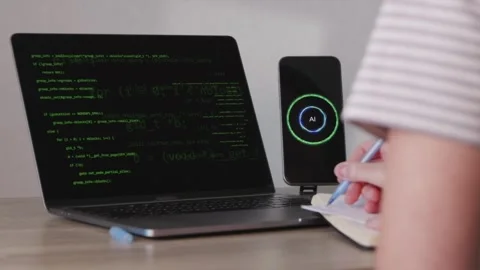 Female developer writing notes next to laptop code and AI phone assistant 스톡 동영상 346834788