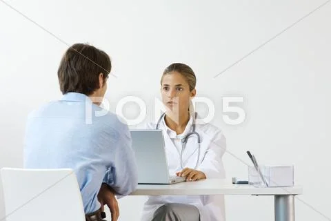Female Doctor Sitting Across From Male Patient At Desk, Using Laptop Computer