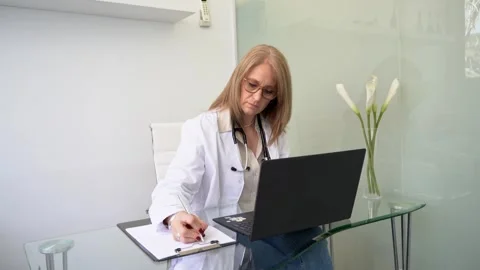 Female doctor taking handwritten notes while reviewing her laptop in the offi Video stock 288048369