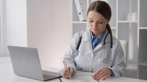 A female doctor writes notes at a modern desk with a laptop. Her role includes Stock Footage 317023492