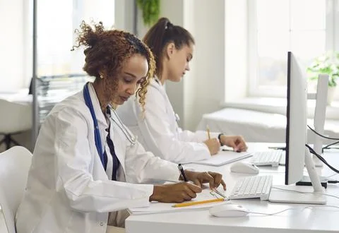 Female doctors analyzing patient data and writing notes at computers in clinic Foto stock