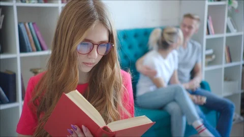 Female dork is reading tutorial while couple hugging on the couch at the library 動画素材 106439662