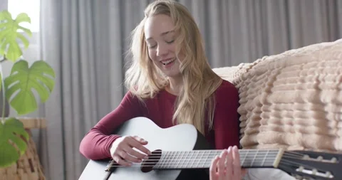 Female early 20s on couch, checking strings, strumming black acoustic guitar, 動画素材 346643186