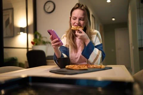 Female eating pizza while using smartphone at kitchen table Stock Photos