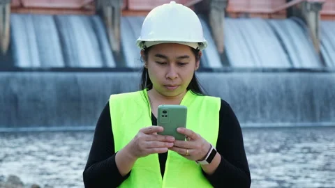 Female engineer in white hat working with smartphone and looking away at dam Stock Footage 229711880