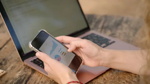 Female freelancer with computer checking statistics charts on smartphone. Woman Stock Footage 239411645