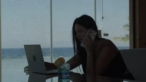 Female freelancer talking on phone while working on laptop in tropical cafe by 스톡 동영상 346250287