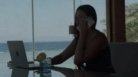 Female freelancer talking on phone while working on laptop in tropical cafe with Stock-Footage 346250880
