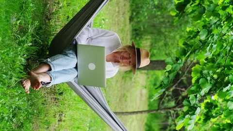 Female freelancer wearing hat using a laptop outdoors. Stockbeeldmateriaal 243633605