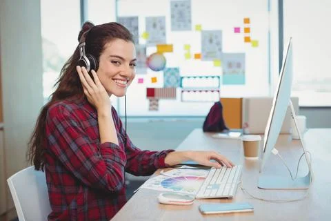 Female graphic designer smiling while adjusting headphones at studio desk with Stock Photos