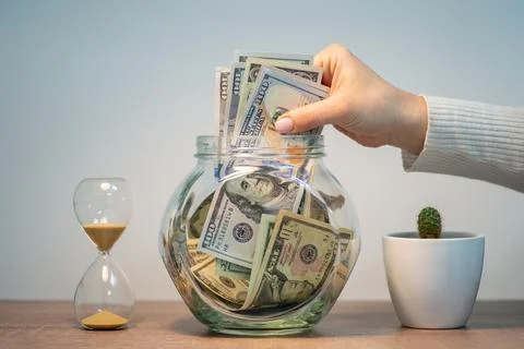 Female hand drops a bunch of dollar banknotes into a glass jar for savings Stock Photos