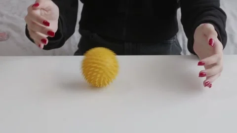 Female hand rolling yellow spiky massage ball on white table surface back forth Stock Footage 319816640