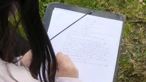 Female hand writing poetry on a paper sheet sitting on the grass. Stock Footage